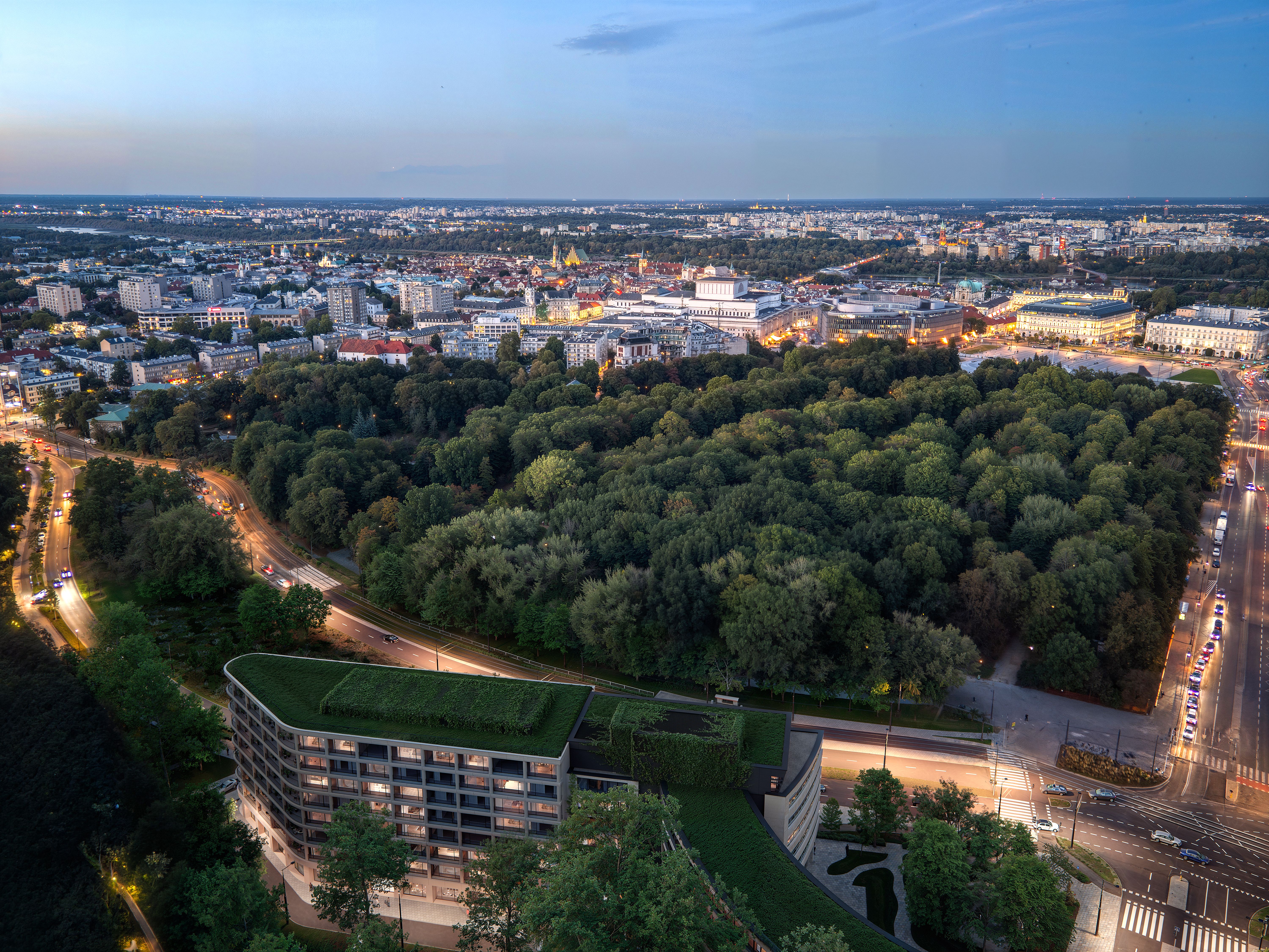 Panoramiczny widok nowoczesnej części miasta w słoneczny dzień. Na pierwszym planie znajduje się szeroka, zadrzewiona aleja z jezdnią i torowiskiem tramwajowym otoczonym bujną zielenią. Po prawej stronie widoczny jest wielopiętrowy budynek mieszkalny z dużymi balkonami i licznymi nasadzeniami roślin. W tle wznoszą się współczesne wieżowce. Nad zabudową rozciąga się bezchmurne, błękitne niebo z pojedynczymi białymi chmurami.